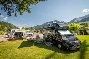 Wohnmobil und Wohnwagen auf einem Campingplatz mit Rasen, Sonnenschirm, Stühlen und Fahrrädern; im Hintergrund Berge und Bäume.