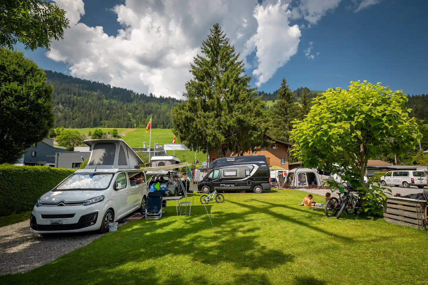 Campingplatz mit Rasenfläche, mehreren Wohnmobilen und Autos, Fahrrädern am Zaun und Bäumen vor grünen Hügeln.