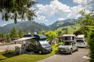 Wohnmobile und Campingbusse auf einem Stellplatz mit Rasen, Markise und Tisch, umgeben von Bäumen und Bergen im Hintergrund.