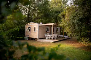 Holz-Mobilheim mit kleiner Terrasse, zwei Stühlen und Tisch vor Bäumen im grünen Campingbereich.