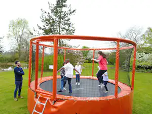 Orangefarbenes Trampolin im Campinggarten, mehrere Personen springen auf dem Netzboden vor Bäumen und Rasen.