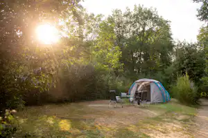 Campingzelt auf einem Stellplatz im Grünen, mit Campingstuhl und Tisch vor dem Zelt, umgeben von Bäumen und Sonnenlicht.