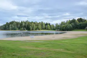 Seeufer mit Sandstrand, Steg und Bootsstegen, umgeben von Rasen und Bäumen unter bewölktem Himmel.
