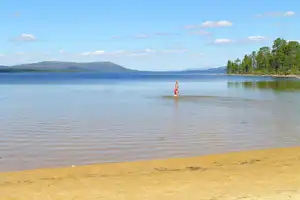 Frau im Wasser am Sandstrand, ruhiger See mit Wald und Hügeln in der Ferne unter blauem Himmel.