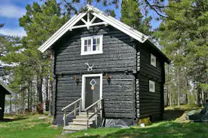 Schwarzes Holzblockhaus mit Eingangstür und Treppe, umgeben von Bäumen im grünen Campingbereich.
