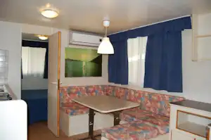 Innenraum eines Wohnwagens mit Sitzbank-Ecke, Tisch, Fenstern und Vorhängen sowie Deckenlampen.