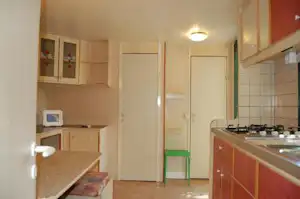 Küchenzeile mit Herd und Spüle in einem Campinghaus, daneben Schrankwand und Tür zum Flur.