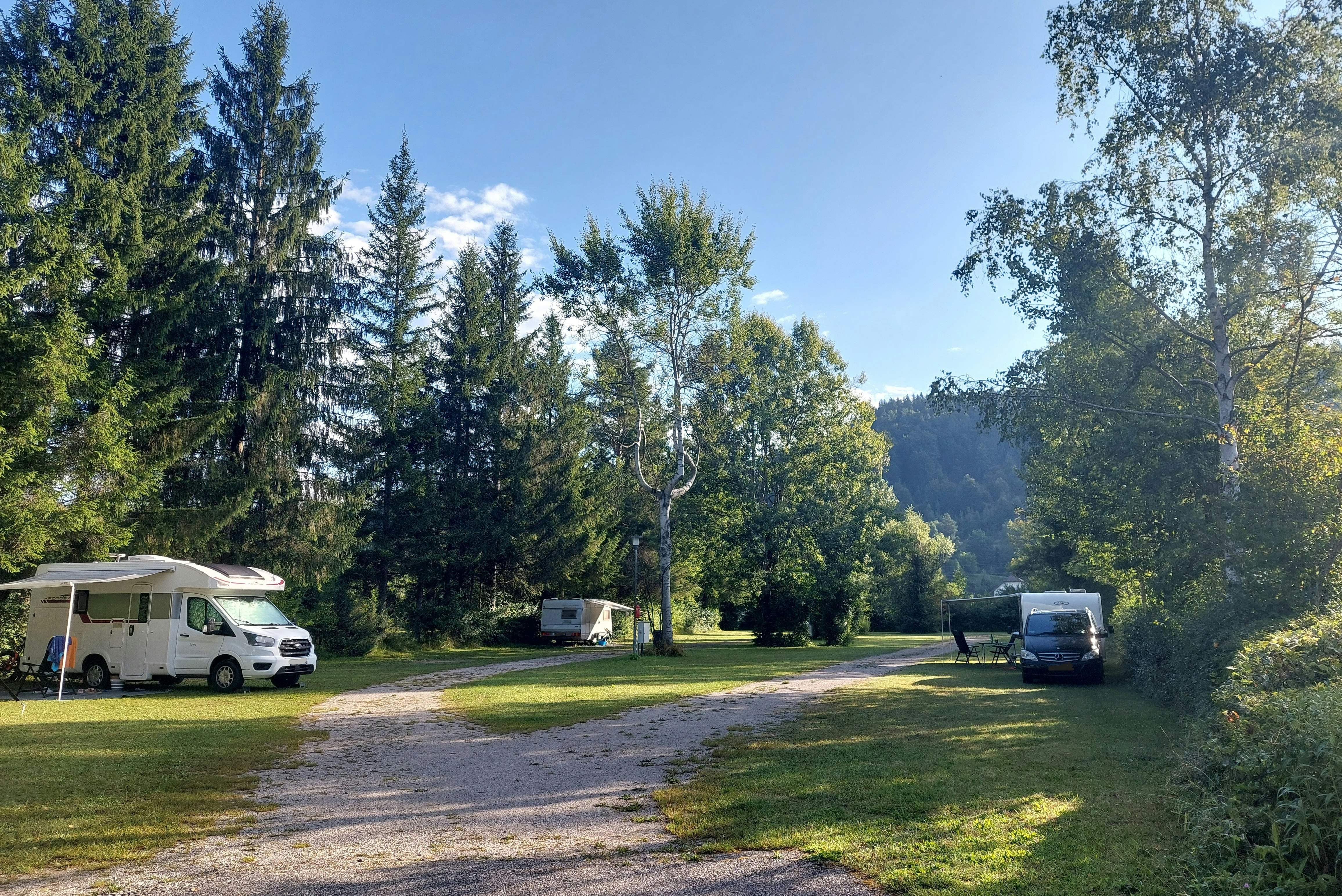 Wohnwagen auf einem Campingplatz in einer Waldlichtung, mit Kiesweg, Rasenflächen und hohen Bäumen im Hintergrund.