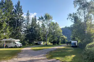 Wohnwagen auf einem Campingplatz in einer Waldlichtung, mit Kiesweg, Rasenflächen und hohen Bäumen im Hintergrund.