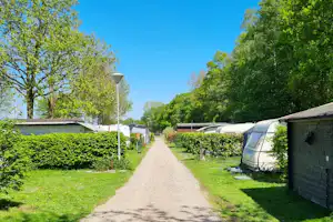 Campingweg zwischen Zelt und Wohnwagen, Hecken und Bäume links, Schotterfläche bis zu einem Gebäude im Hintergrund.