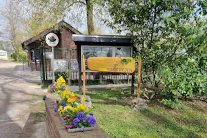 Blumenbeet am Weg zum Camping-Eingang mit Ziegelhaus, Schildern und Bäumen im Hintergrund.