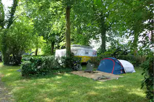 Wohnmobil und Zelt auf einem Stellplatz im Grünen unter Bäumen, mit Fahrrad und Campingstuhl neben der Markise.