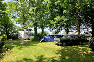 Wohnmobil und blaues Zelt auf einem grünen Campingplatz, daneben ein Oldtimer und weitere Fahrzeuge unter Bäumen.