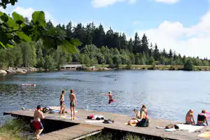 Holzsteg am See mit Badegästen, im Hintergrund Wald und ein kleiner Steg am Ufer mit Steinen.