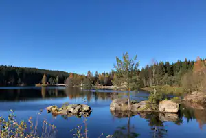 Ruhiger See mit Spiegelung, Steg und Uferfelsen, umgeben von Wald und einzelnen Bäumen unter klarem Himmel.