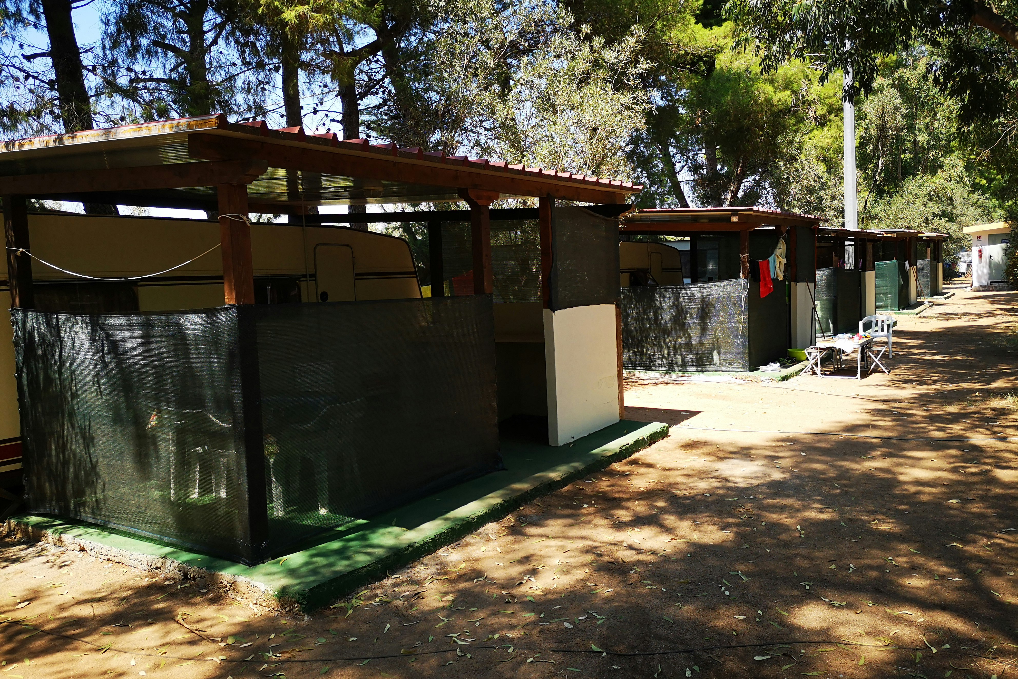 Carport am Campingplatz mit Sichtschutzwand, Stellplatz für Wohnmobil und Tisch im Schatten von Bäumen.