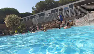 Blaues Camping-Poolbecken im Vordergrund, dahinter Terrassen mit Geländern und Bungalows, umgeben von Bäumen und Hecken.
