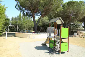 Spielplatz am Campingplatz mit grünem Kletterturm, Sandfläche, Schaukel und Bäumen im Hintergrund.