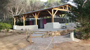 Holz-Pavillon mit Sitzbänken auf einer erhöhten Plattform, umgeben von Bäumen und Kiesweg am Campingplatz.