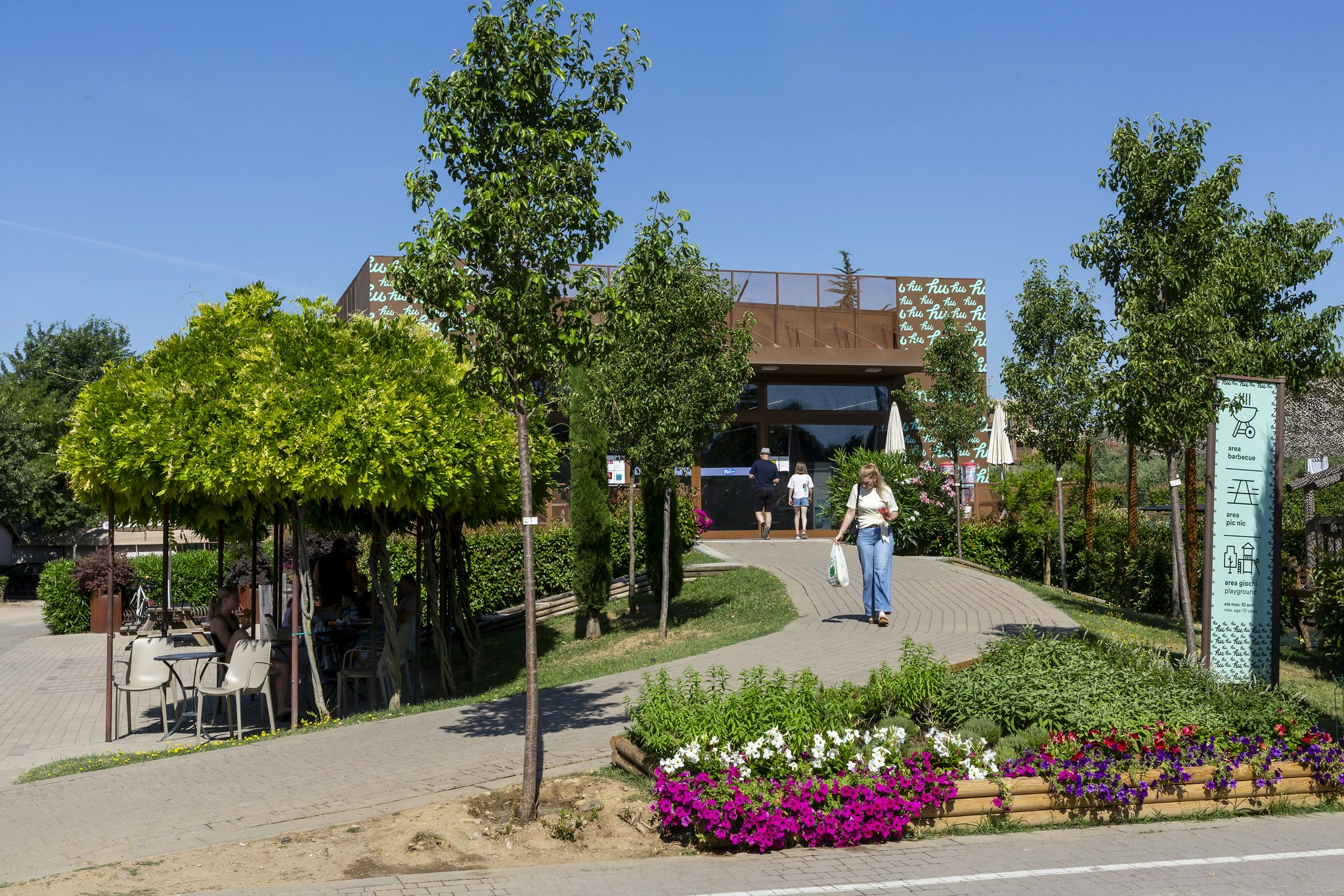 Eingang eines Campingplatzes mit Weg, Blumenbeeten und Bäumen, im Hintergrund ein Gebäude mit Holzüberdachung.