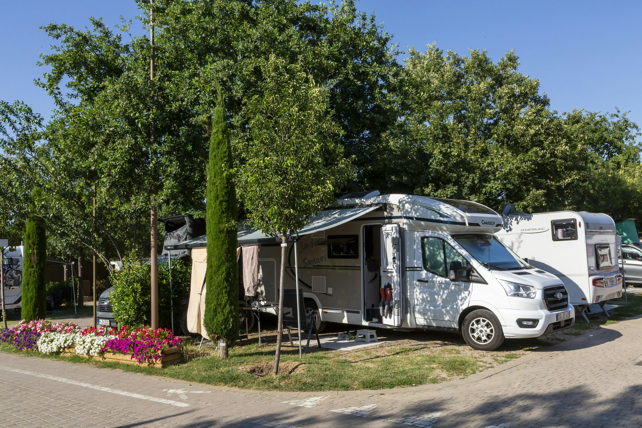 Wohnmobil und Caravan auf einem Campingplatz, neben Hecken und Blumenbeeten, mit Stellplatz am Weg und Bäumen im Hintergrund.