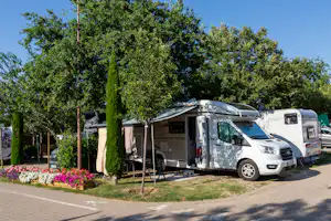 Wohnmobil und Caravan auf einem Campingplatz, neben Hecken und Blumenbeeten, mit Stellplatz am Weg und Bäumen im Hintergrund.