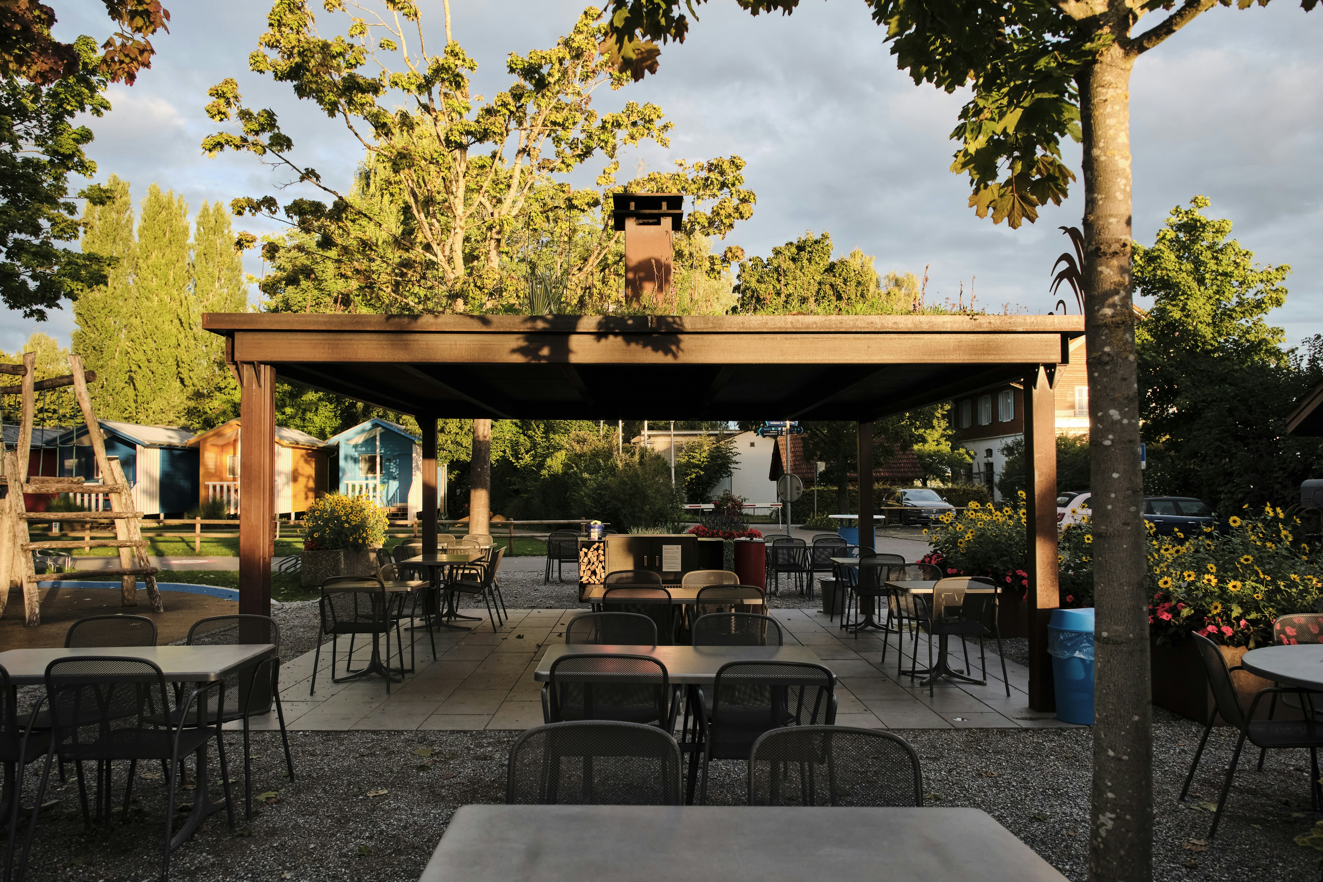 Überdachte Terrasse mit vielen Stühlen und Tischen, im Hintergrund Bäume und ein Gebäude am Campingplatz.