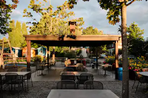 Überdachte Terrasse mit vielen Stühlen und Tischen, im Hintergrund Bäume und ein Gebäude am Campingplatz.