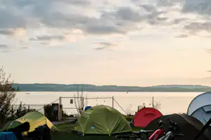 Zelte auf einem Campingplatz am Seeufer, mit Steg und Wasser im Hintergrund, im Vordergrund Rasen und Campingausrüstung.