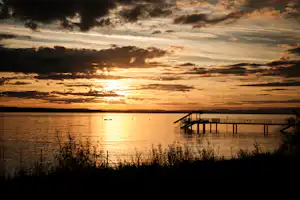 Sonnenuntergang über einem See mit Steg und Ufervegetation, im Hintergrund dunkle Wolken und warmes Licht.