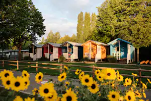 Bunte Camping-Häuschen mit Veranda und Holztreppe, im Vordergrund gelbe Sonnenblumen vor grünen Bäumen und Rasen.