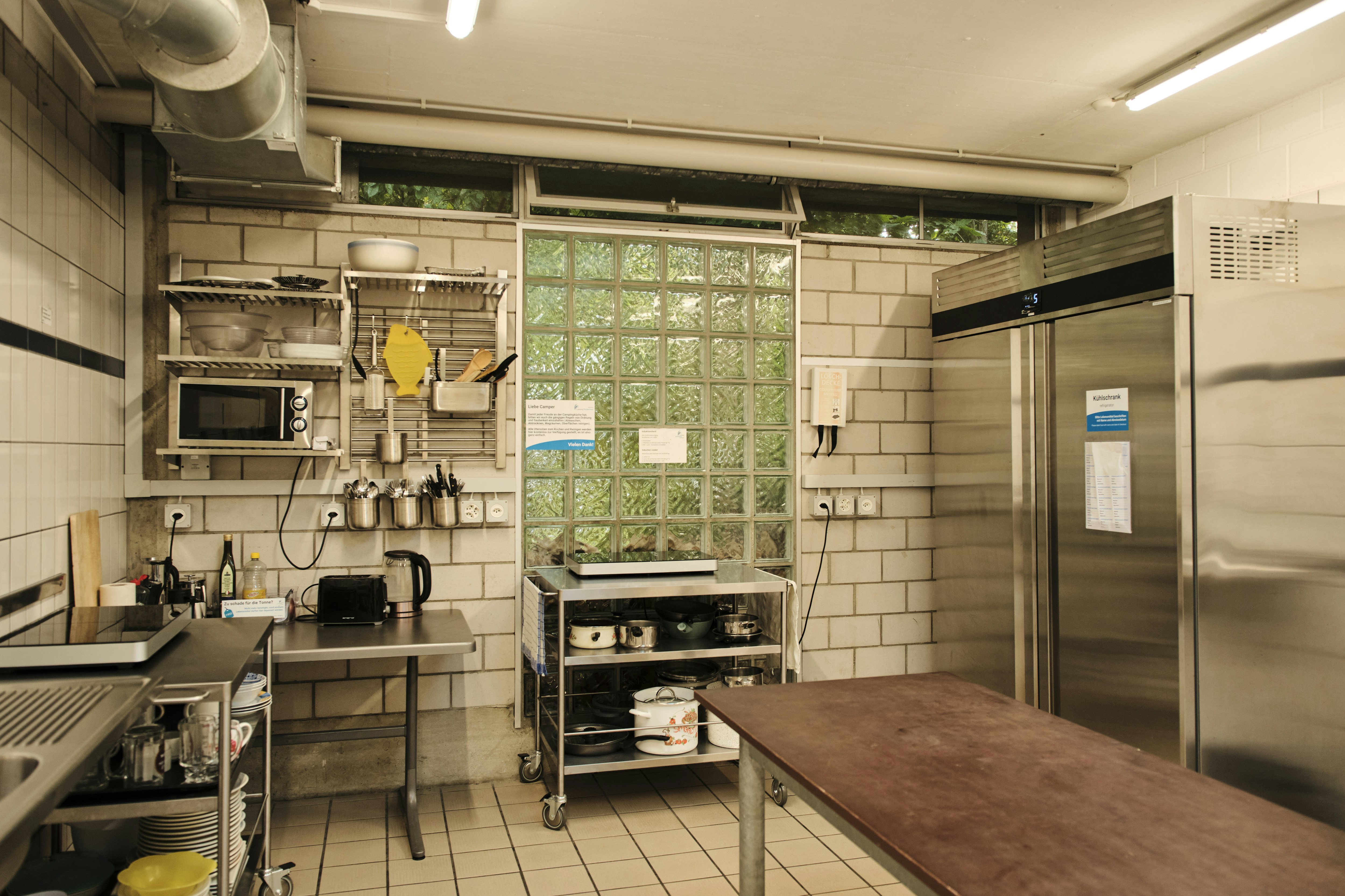 Gemeinschaftsküche mit Spüle, Herd und Arbeitsflächen, daneben ein Grill und ein großer Kühlschrank, im Hintergrund Fenster mit Gitter.