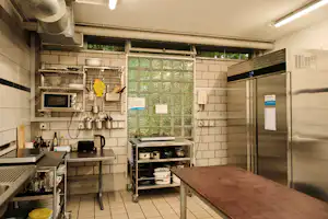 Gemeinschaftsküche mit Spüle, Herd und Arbeitsflächen, daneben ein Grill und ein großer Kühlschrank, im Hintergrund Fenster mit Gitter.
