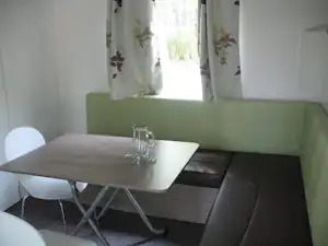 Essbereich im Wohnwagen: grauer Tisch mit Glaskrug und weißen Stühlen, grüne Sitzbank und Fenster mit Vorhängen.