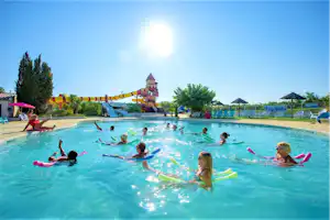 Kinder im Schwimmbecken eines Campingplatzes, mit Rutschen und Spielturm im Hintergrund, Sonnenschein am Himmel.