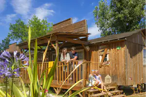 Holzterrasse am Campinggebäude mit zwei Personen, Blumenbeeten und Holzgeländer unter blauem Himmel.