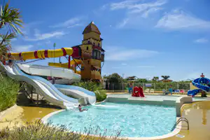 Großer Camping-Pool mit Wasserrutsche, buntem Spielturm und Liegen am Beckenrand, im Hintergrund Palmen und Gebäude.