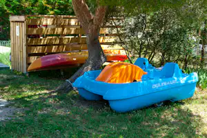 Aufblasbares Boot in Blau und Orange vor einem Holzsichtschutz, im Schatten eines großen Baums im Campingbereich.