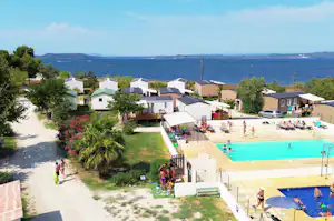 Campingplatz mit Pool und Liegewiese, Reihen von Wohnwagen und kleinen Häusern am Ufer, Blick auf das Wasser.