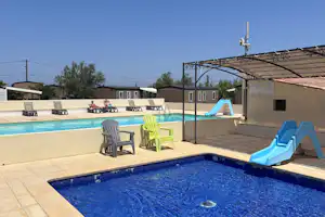 Blauer Swimmingpool mit Rutsche, Liegestühlen und Sonnenschirm am Campingplatz, daneben Sitzbereich und Gebäude im Hintergrund.