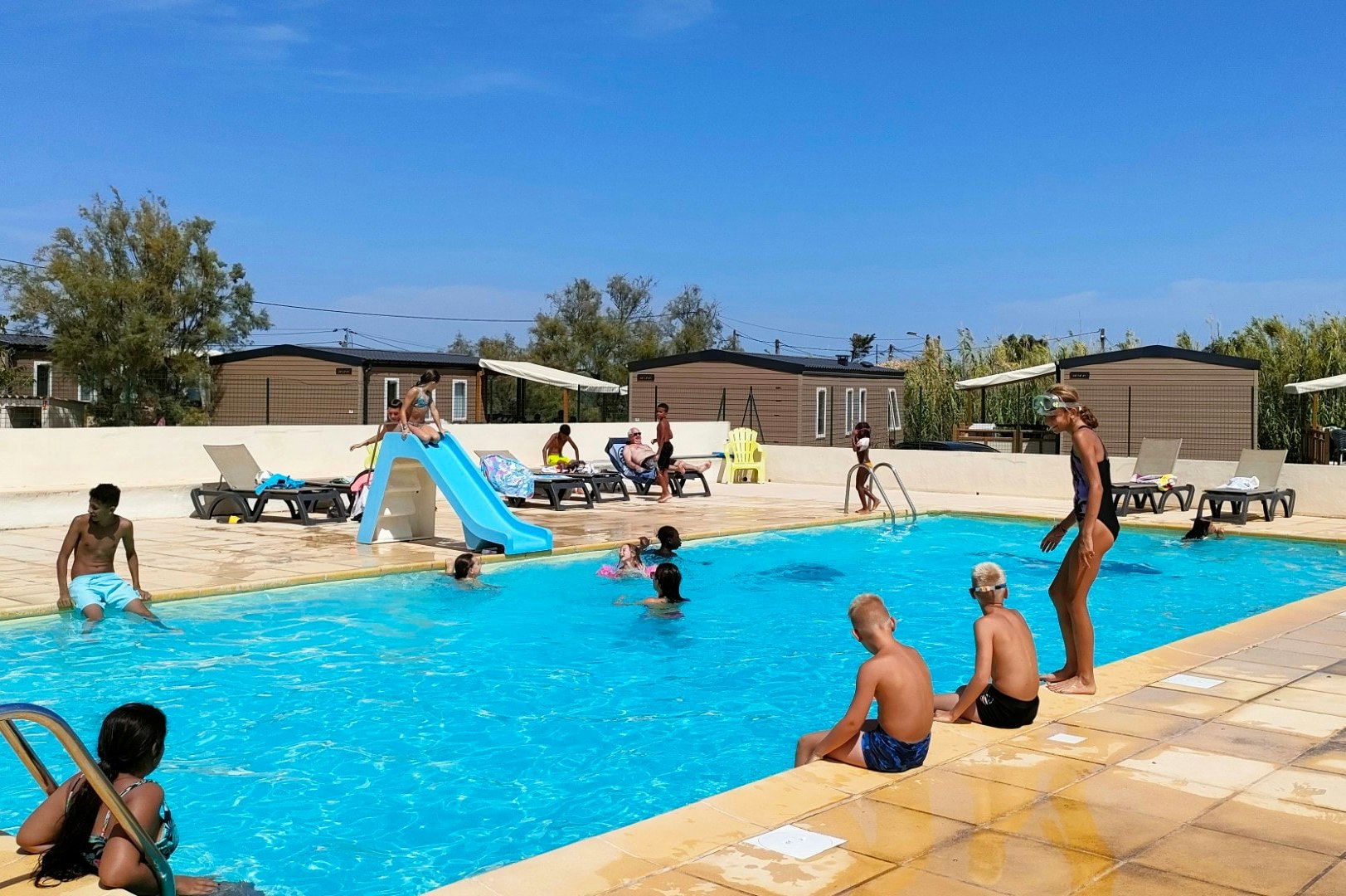 Kinder und Erwachsene im Camping-Pool mit Rutsche, Liegen und Sonnenschirmen auf der Terrasse, im Hintergrund Wohnwagen und Gebäude.