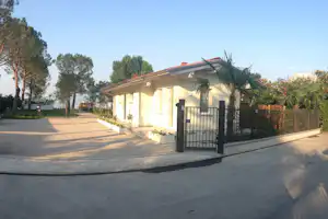 Campinghaus mit Einfahrt und schwarzem Tor, gepflasterter Weg, Zaun und Bäume am Rand des Geländes.