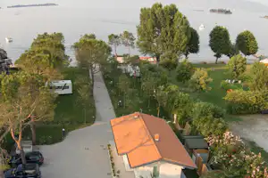 Blick auf einen Campingplatz am See mit Wohnwagen, gepflastertem Weg, Bäumen und einem kleinen Gebäude am Ufer.
