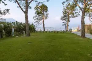 Grüner Campingrasen mit Bäumen, Kiesweg und Blick auf den See mit Steg und Booten in der Ferne.