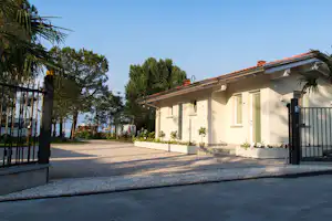 Campinghaus mit rotem Ziegeldach, Einfahrt und schwarzem Tor, daneben Bäume und Palmen am Weg.