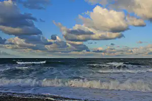 Wellen schlagen an der Küste auf, dunkle und helle Wolken am Himmel über dem Meer.