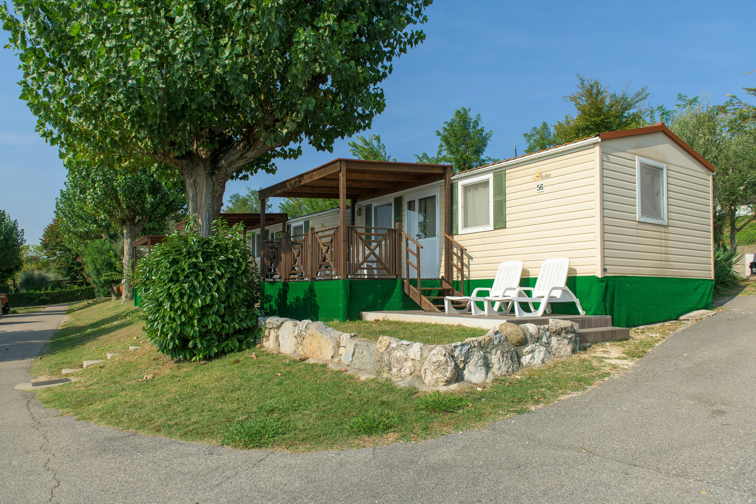 Mobilheim mit Holzterrasse, zwei Liegestühlen und grüner Umrandung, umgeben von Bäumen und Rasen am Campingplatz.