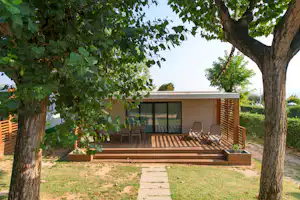 Bungalow mit Terrasse, Holzgeländer und Liegestühlen, umgeben von Bäumen und Grün im Campingbereich.