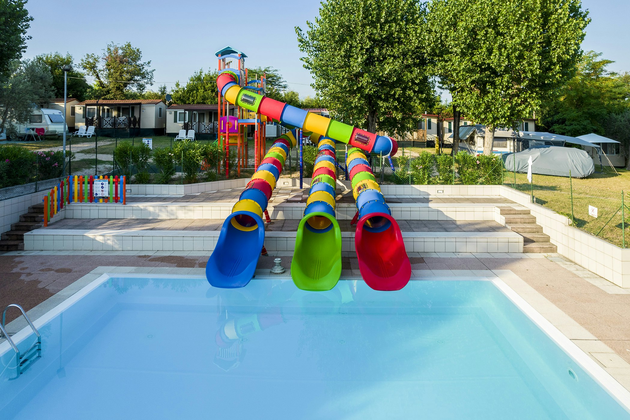 Kinderwasserrutsche am Campingplatz neben dem Pool, mit bunten Rutschen und Liegen, Bäumen und Zelten im Hintergrund.