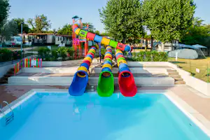 Kinderwasserrutsche am Campingplatz neben dem Pool, mit bunten Rutschen und Liegen, Bäumen und Zelten im Hintergrund.